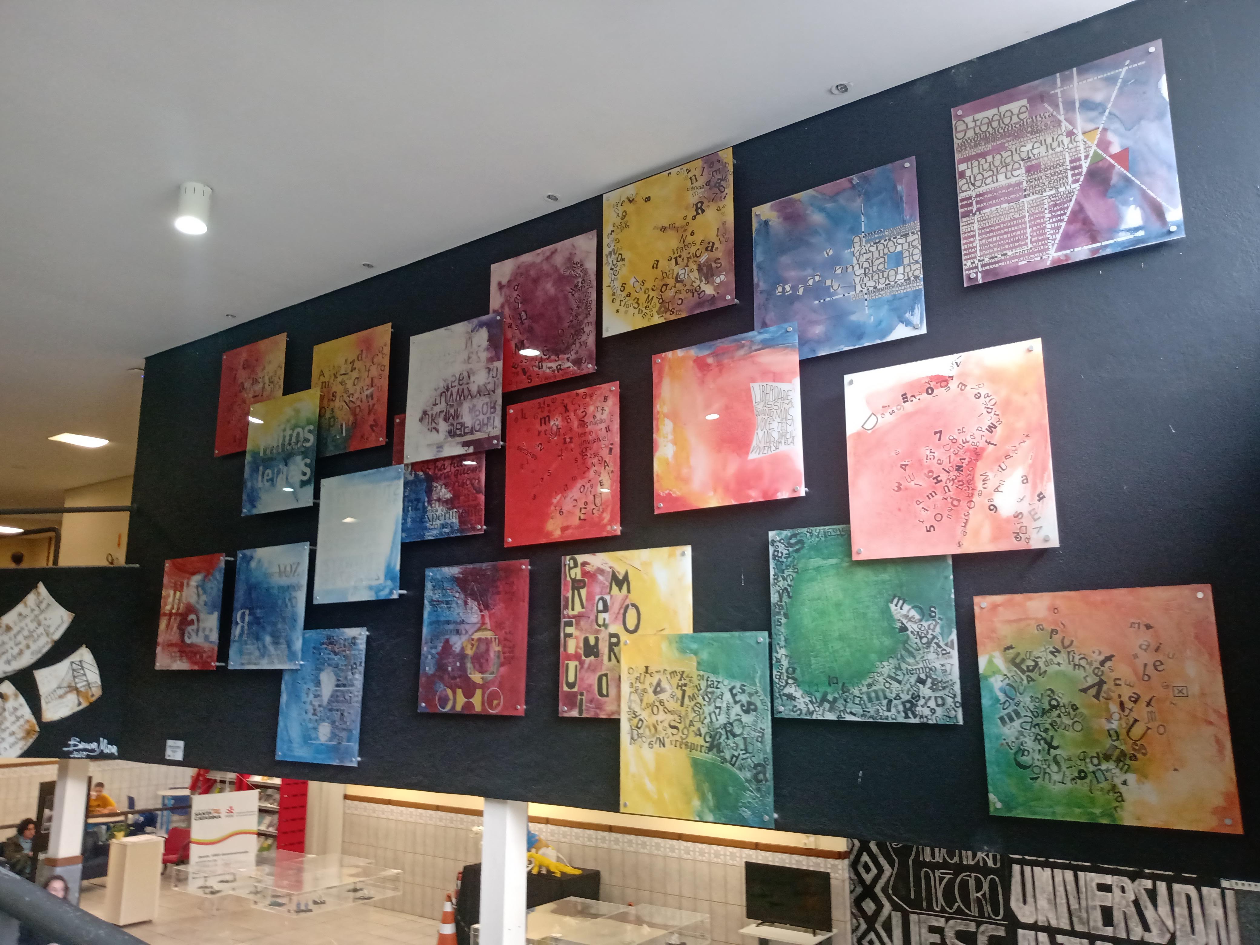 Foto colorida em diagonal mostrando uma parede preta com várias pinturas multicoloridas. Na parte inferior da fotografia é possível ver parte do hall de entrada da biblioteca da UFSC.