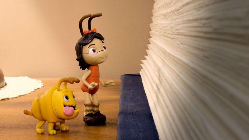 Cena do filme Teca e Tuti: uma noite na biblioteca. Na imagem aparece a traça Teca (tronco vermelho, sapato marrom e laço na cabeça) e o ácaro Tuti (corpo amarelo e com língua de fora) olhando para a lateral de um livro.